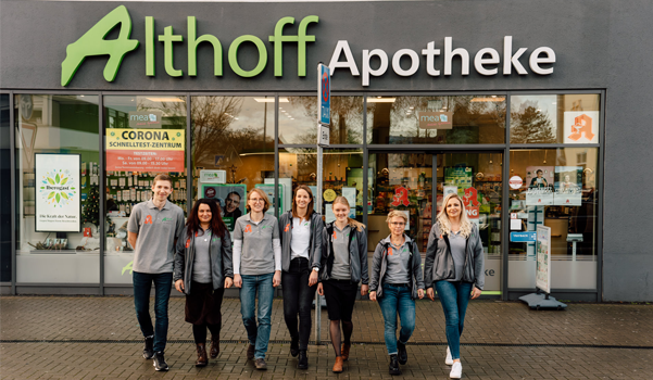 Team der Althoff Apotheke steht vor dem Eingangsbereich der Apotheke in Neuwied, Glasfront mit Apothekenlogo und Hinweisschildern.